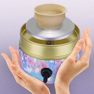 Op de afbeelding: Een klein pottenbakkerswiel met een bloemenpatroon. Het wiel is goudkleurig met een blauw en roze bloemenpatroon. Een kleine klei kom staat op het wiel. Het wiel heeft een knop om de snelheid te regelen.