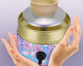 Può includere: Un piccolo tornio da vasaio con un design floreale. Il tornio è dorato con un motivo floreale blu e rosa. Una piccola ciotola di argilla è sul tornio. Il tornio ha una manopola per controllare la velocità.