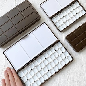 Puede incluir: Varias paletas de acuarela marrones con pozos de pintura blancos están abiertas y cerradas sobre una superficie blanca. Las paletas tienen forma rectangular con tapa y una sección para mezclar colores. Una paleta está abierta.