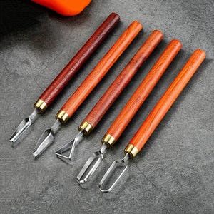 May include: Five wood-handled carving tools with stainless steel blades. The handles are various shades of brown and orange, with brass-colored metal accents. The blades have different shapes, including a V-shape and a rounded edge. The tools are arranged diagonally.