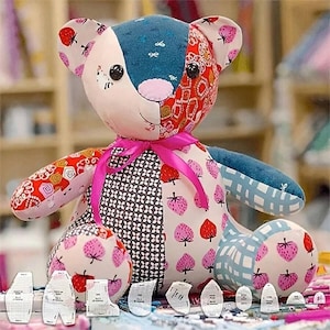 May include: A patchwork teddy bear with a pink ribbon around its neck. The bear is made of different fabrics with patterns of strawberries, flowers, and geometric designs. The bear is sitting on a white surface with several paper patterns.