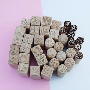 Può includere: Una collezione di timbri in legno con vari disegni intagliati, tra cui fiori, motivi geometrici e simboli. I timbri hanno forme diverse, alcuni quadrati e altri cilindrici, e sono realizzati in legno chiaro.
