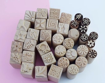 May include: A collection of wooden stamps with various carved designs, including flowers, geometric patterns, and symbols. The stamps are in different shapes, some square and some cylindrical, and are made of light-colored wood.