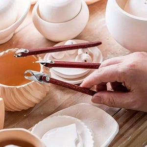 May include: A selection of pottery tools, including wooden-handled trimming tools with metal blades, are arranged near ceramic bowls and plates. The tools are being held near a ceramic bowl with a textured, shell-like design. The scene suggests a pottery studio.