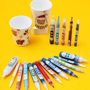 May include: A collection of colorful paint pens and two white ceramic mugs with festive designs on a yellow background. The pens are labeled with various colors and Chinese characters. The mugs feature holiday-themed illustrations.