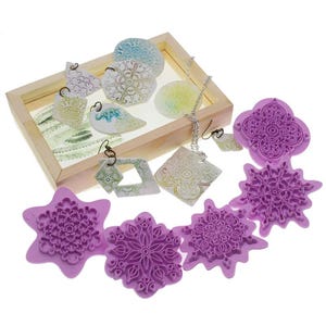 Può includere: Una collezione di gioielli fatti a mano e stampi in argilla polimerica. I gioielli includono orecchini e una collana con motivi intricati in bianco, verde e blu. Sono presenti anche stampi viola a forma di fiocco di neve e floreali.