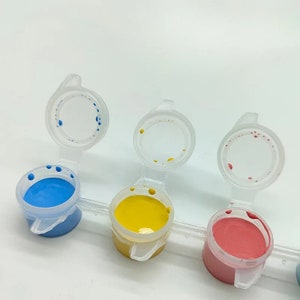 K&ouml;nnte beinhalten: Eine Reihe von Farbt&ouml;pfen mit blauer, gelber und roter Farbe. Jeder Topf hat einen transparenten Klappdeckel. Die Farbe ist in den offenen T&ouml;pfen sichtbar, mit einigen Farbtropfen auf den Deckeln. Die T&ouml;pfe sind in einer Reihe angeordnet.