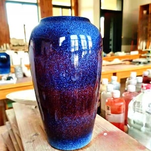 Puede incluir: Un jarrón de cerámica cilíndrico con un esmalte degradado que transita de un azul oscuro profundo en la parte superior a un rico marrón rojizo en la base. La superficie tiene una apariencia moteada y texturizada con un acabado brillante.