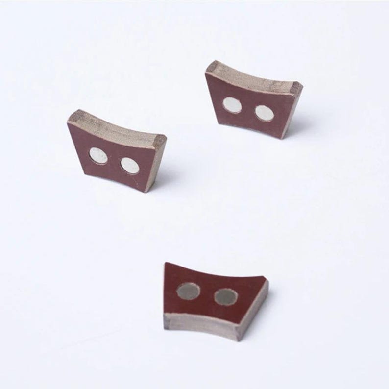 Pu&ograve; includere: Tre piccoli oggetti curvi a forma di trapezio. Ogni oggetto ha una faccia rosso scuro con due inserti circolari color argento. I bordi sono di un colore pi&ugrave; chiaro e neutro. Gli oggetti sono disposti su uno sfondo bianco.