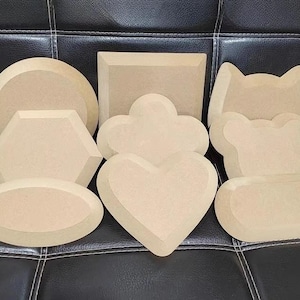May include: A collection of unfinished wooden shapes in various forms, including a circle, square, hexagon, oval, cloud, heart, and a cat-like design. The shapes are light brown and appear to be made of MDF or similar material.