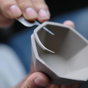 May include: A light grey, octagonal-shaped silicone mould being trimmed with a small metal tool. The mould has a smooth, matte finish and a distinct geometric rim. The tool is positioned along the top edge to remove excess material from the form.