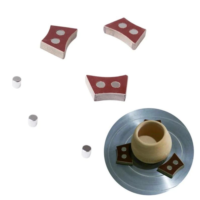 Pu&ograve; includere: Una collezione di strumenti per la ceramica, tra cui diversi magneti rossi e grigi e piccoli cilindri argentati. Gli strumenti sono disposti attorno a un piccolo vaso di ceramica marrone chiaro su una superficie metallica argentata.