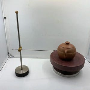 以下が含まれることがあります： 蓋付きの茶色の陶器製ティーポットが円形の回転台の上に置かれています。垂直の棒と水平の針アームを備えた金属製のスタンドがティーポットの横に配置されており、針の先端が陶器の側面に触れています。