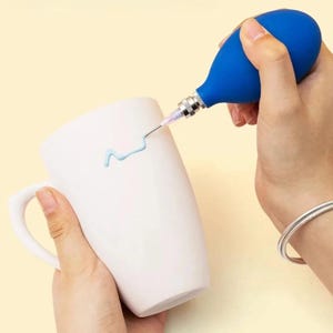 Puede incluir: Una taza de cerámica blanca con asa negra y el texto "But First, Coffee" en negrita negra. La taza se encuentra sobre un fondo borroso, sugiriendo un ambiente acogedor.