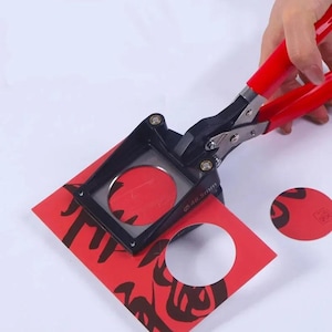 May include: A red paper circle cutter with black and silver parts. The cutter is over a red square of paper with black calligraphy. The cutter has a 48.5mm diameter. A hand holds the red handles.
