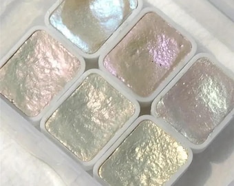 May include: A set of six pearlescent watercolor paints in square white plastic containers. The colors range from pale gold to iridescent pink and blue, with a shimmering, textured surface. The paints are arranged in a clear plastic tray.