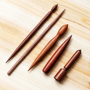 May include: Five handcrafted wooden tools in varying shapes and sizes, displayed on a light wood surface. The tools are a rich, dark brown color, with smooth, polished finishes. The tools appear to be for crafting or woodworking.