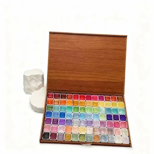 May include: A wooden box with a hinged lid, open to reveal a collection of 72 watercolor paints in a rainbow of colors. The paints are arranged in neat rows, each in its own square well. The box is made of a light brown wood.