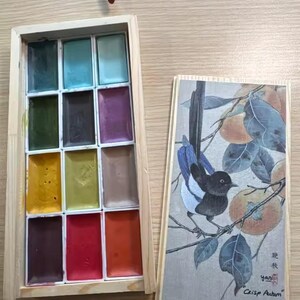 May include: A wooden box of watercolor paints with a variety of colors, including blues, greens, yellows, reds, and browns. Next to it is a wooden box with a painting of a bird on a branch with the text "Crisp Autumn" and "Chinese painting Pigments".