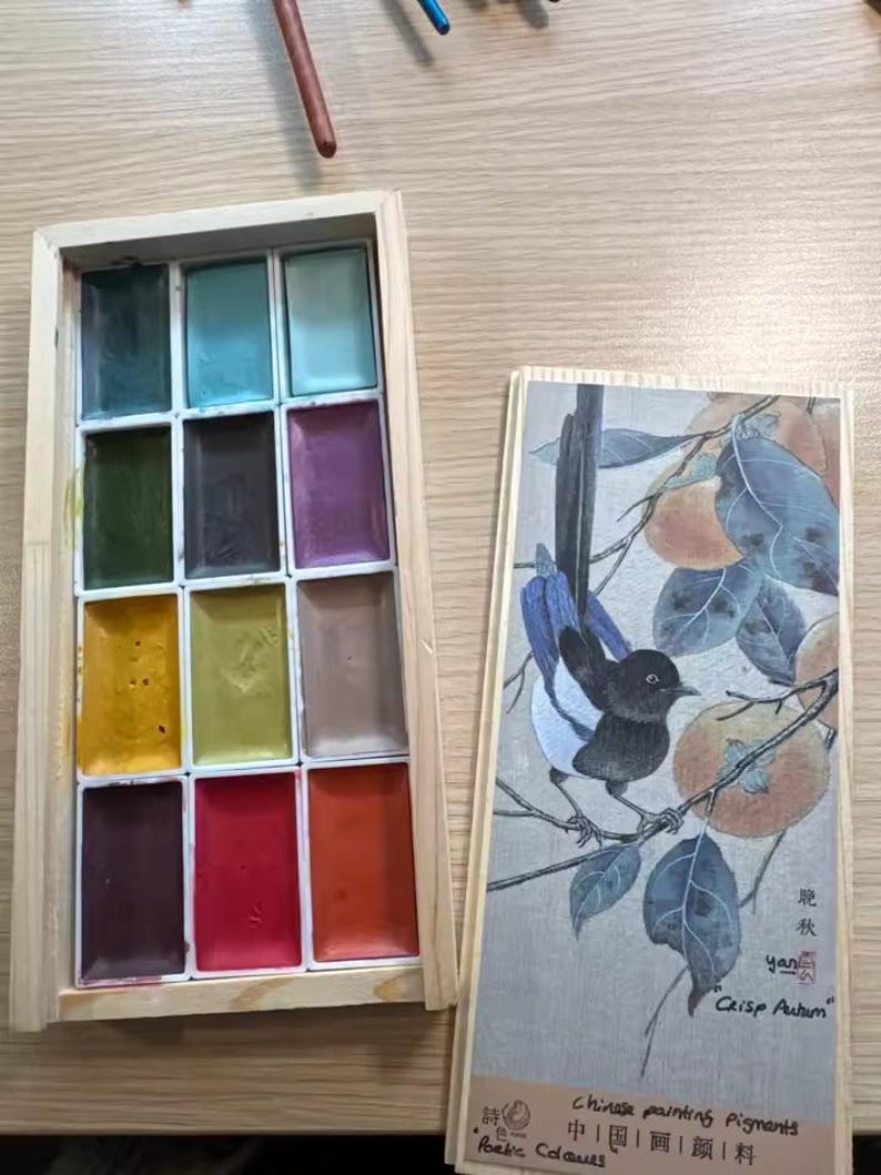 May include: A wooden box of watercolor paints with a variety of colors, including blues, greens, yellows, reds, and browns. Next to it is a wooden box with a painting of a bird on a branch with the text "Crisp Autumn" and "Chinese painting Pigments".