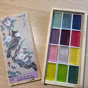 May include: A wooden box set of Chinese painting pigments with 12 vibrant colors. The lid features a detailed illustration of a bird on a flowering branch, with Chinese text and the words "Easy Spring".