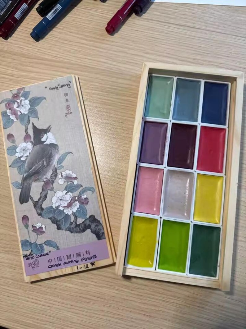 May include: A wooden box set of Chinese painting pigments with 12 vibrant colors. The lid features a detailed illustration of a bird on a flowering branch, with Chinese text and the words "Easy Spring".