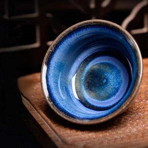 Puede incluir: Una pequeña taza de té de cerámica con un interior azul profundo y un borde marrón oscuro. El interior de la taza presenta un patrón arremolinado de azules y verdes, creando un diseño artístico único. La taza está colocada sobre una superficie de madera.