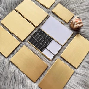 Peut inclure: Plusieurs étuis rectangulaires dorés, certains ouverts révélant un intérieur noir et blanc. Les étuis sont disposés sur une surface douce et grise. Une petite figurine est également présente.