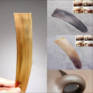 May include: A close-up of a brown, curved, horn tea scraper. The scraper is made of natural horn and has a smooth, polished surface. The scraper is used to clean the inside of teapots and cups.