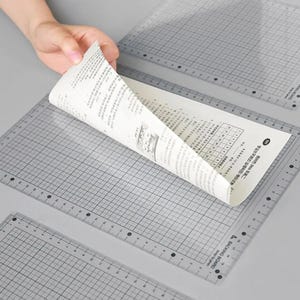 Puede incluir: Alfombrillas de corte transparentes con cuadrícula y bordes de regla. Una mano levanta una hoja de papel, revelando texto y una tabla. Las alfombrillas están etiquetadas como "BACKING BOARD" y tienen medidas en cm y pulgadas.