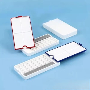May include: Three rectangular white plastic watercolour paint boxes with hinged lids. One box has a red border, one has a blue border, and one is closed. Each open box features a grid of small square wells for paint and a central mixing area.