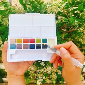 May include: A white watercolor paint palette with an assortment of colorful paints. A clear water brush is held over the palette. The palette is open, revealing various shades of paint, including blue, green, red, and yellow.