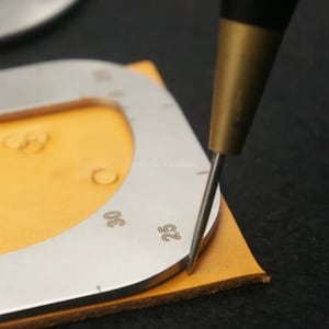 May include: A silver metal tool with a circular cutout and markings for different sizes. The tool is placed on a piece of tan leather and a small awl is being used to mark the leather.