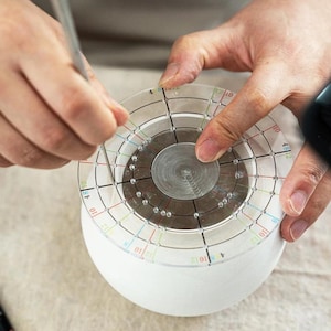 Puede incluir: Una persona utiliza una herramienta circular transparente con líneas y números de colores para medir un objeto esférico blanco. La herramienta está centrada en el objeto y se sujeta con las manos de la persona. La herramienta tiene un centro de metal.