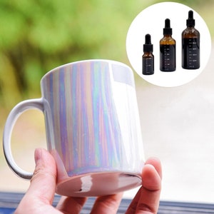 以下が含まれることがあります： 持ち手のついた白い虹色のセラミックマグカップ。マグカップは手で持たれています。背景には、ドロッパー付きのガラス瓶が3本あります。瓶は暗い液体で満たされており、側面にミリリットルを示すマークが付いています。