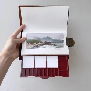 May include: A watercolor painting of a coastal scene is displayed in a brown leather-bound sketchbook. The sketchbook has a built-in red paint palette with white mixing areas. The painting depicts a beach, ocean, and mountains.