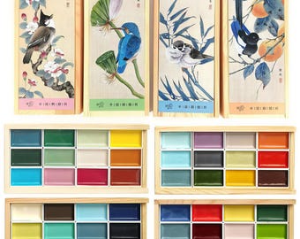 May include: Watercolor paint sets in wooden boxes. Each set features 16 rectangular paint pans in various colors. The top row of each box has a different illustration of a bird and floral design. The sets are in a variety of colors.
