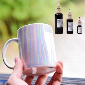 Peut inclure: Une tasse en céramique blanche avec une finition arc-en-ciel irisée. Trois petits flacons de liquide foncé avec des compte-gouttes sont à l'arrière-plan.