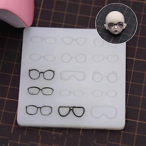 May include: A white silicone mold with various eyeglasses designs. The mold includes several styles of glasses, including round, oval, and cat-eye shapes. A doll head with glasses is in the upper right corner.