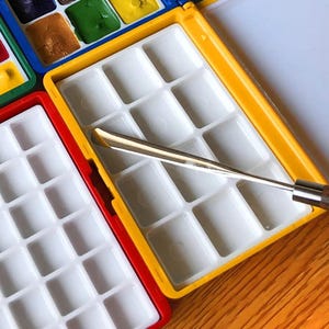 May include: A close-up of open watercolor palettes with various colors of paint. The palettes are red, yellow, and blue. A silver and wood-handled palette knife rests on the yellow palette. The paint colors include brown, green, and yellow.