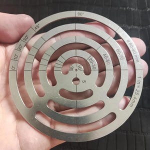 May include: A silver metal circle with concentric rings marked with measurements in inches and millimeters. The outer ring is marked with measurements from 60-70, 40-50, 20-30, and 10. The inner ring is marked with measurements from 0-10, 20, 30, 45, and 90 degrees. The text "1 in = 25.4 mm" and "d 80-90 mm" is printed on the circle.