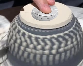 May include: A pottery wheel with a ceramic bowl being shaped on it. A white ceramic disc is on top of the bowl, and a hand is pressing down on the disc.