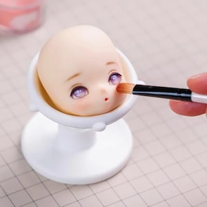 Puede incluir: Una cabeza de muñeca con ojos morados y mejillas rosadas se sostiene en un soporte blanco. Un pequeño pincel aplica maquillaje en la cara de la muñeca. La cabeza de la muñeca es pálida con rasgos delicados.