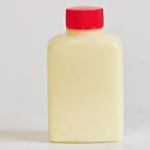 Op de afbeelding: Een kleine, rechthoekige, ondoorzichtige gebroken witte plastic fles met afgeronde hoeken en een geribbelde, felrode schroefdop. De container heeft een gladde, matte afwerking en een platte bodem, ontworpen voor het rechtop bewaren van vloeistoffen.