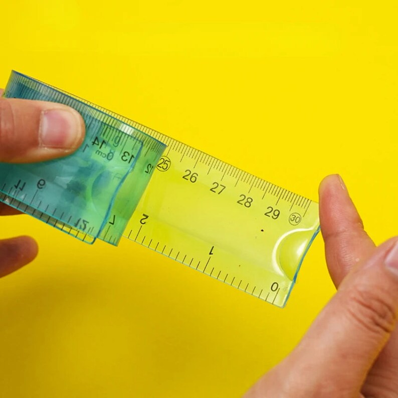 May include: A green plastic ruler with a clear plastic overlay. The ruler has markings for inches and centimeters. The overlay has markings for centimeters only. The ruler is being held by a hand.