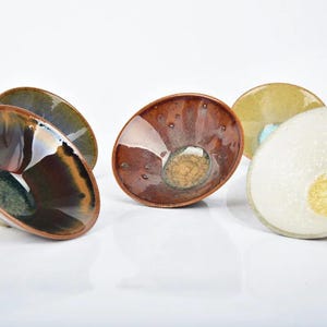 Puede incluir: Colección de cuencos de cerámica hechos a mano con formas orgánicas únicas. Los cuencos presentan una variedad de esmaltes en tonos tierra, incluyendo tonos de marrón, verde y crema, con acabados brillantes y texturizados. Los cuencos se exhiben sobre una superficie blanca.