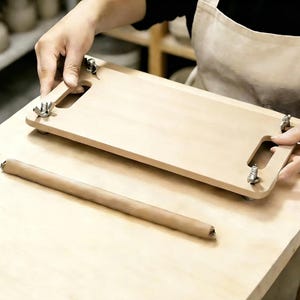 Può includere: Un utensile rettangolare in legno chiaro con due manici ritagliati e quattro bulloni filettati in metallo con dadi a farfalla agli angoli. Un lungo e sottile pezzo cilindrico di argilla marrone giace parallelo all'utensile su una superficie.