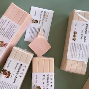 Può includere: Sono esposti diversi pacchi di argilla polimerica in varie tonalità di rosa e beige. Ogni confezione è etichettata con testo in lingua straniera e le parole "Polymer Clay". È visibile anche un piccolo strumento di argilla rosa.