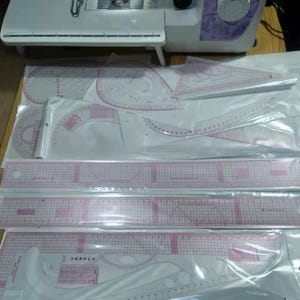 May include: A collection of clear plastic sewing rulers and templates with pink grid lines and markings. Various shapes include straight rulers, curves, and protractors. A white sewing machine is visible in the background.