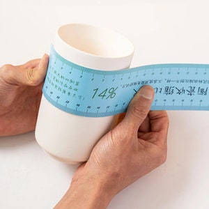 May include: A blue measuring tape with a white ceramic mug. The tape reads "Ceramic Shrinkage Ratio" and "14%".